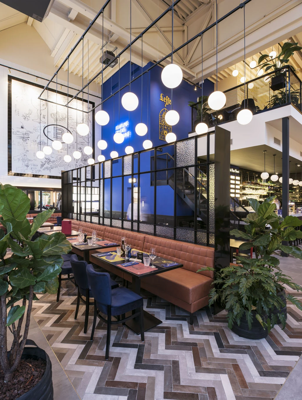 The double-height dining room at Le Petit Belge Maasmechelen Village, with globe lights, the blue wall, leather banquettes and a herringbone floor